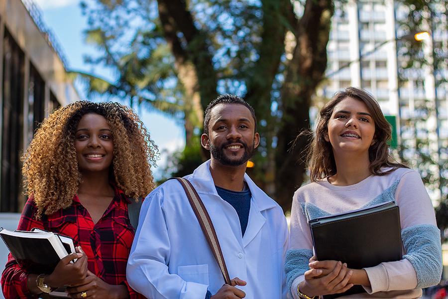 Three students in a row smiling, two students holding textbooks, and one student holding backpack strap.